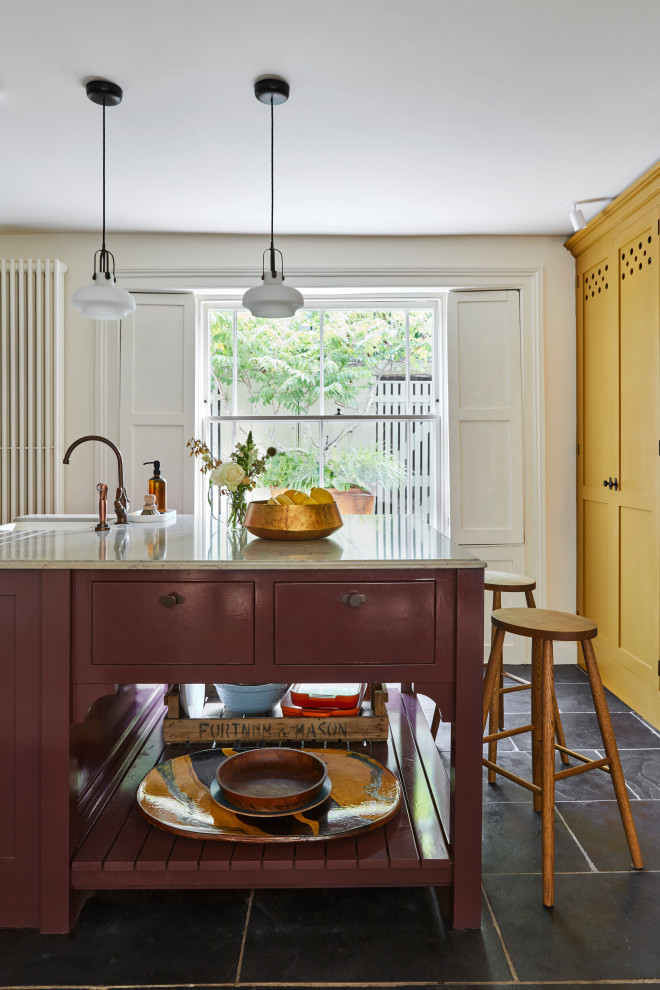 This is an example of a large transitional eat-in kitchen in London with a farmhouse sink, shaker cabinets, yellow cabinets, marble benchtops, white splashback, ceramic splashback, white appliances, limestone floors, with island, black floor and brown benchtop.
