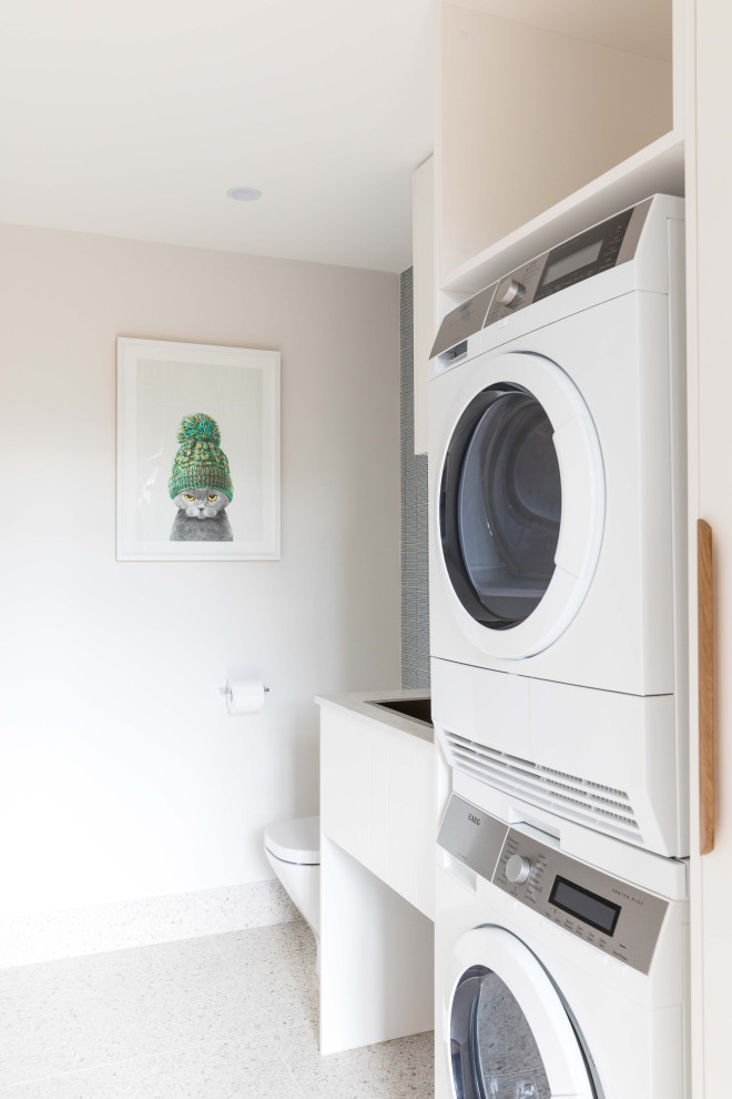 Design ideas for a small midcentury single-wall dedicated laundry room in Sydney with a drop-in sink, white cabinets, quartz benchtops, white walls, ceramic floors, multi-coloured floor, white benchtop and a stacked washer and dryer.