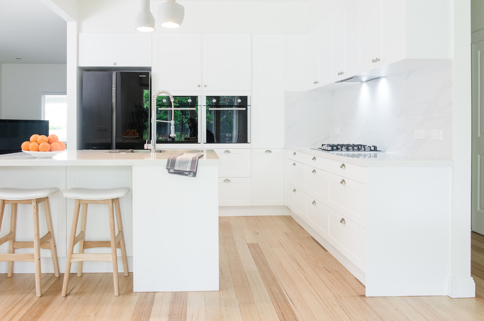 Inspiration for a large scandinavian l-shaped kitchen pantry in Melbourne with a double-bowl sink, shaker cabinets, white cabinets, solid surface benchtops, white splashback, ceramic splashback, black appliances, light hardwood floors and with island.