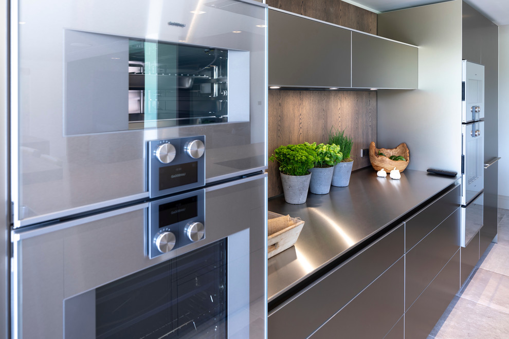 Inspiration for a large contemporary open plan kitchen in Cheshire with an integrated sink, flat-panel cabinets, medium wood cabinets, stainless steel benchtops, timber splashback, stainless steel appliances, ceramic floors, multiple islands, white floor and green benchtop.