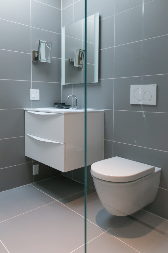 Modern inredning av ett vit vitt badrum, med vita skåp, en kantlös dusch, en vägghängd toalettstol, grå kakel, porslinskakel, grå väggar, klinkergolv i porslin, ett väggmonterat handfat, bänkskiva i glas, grått golv och med dusch som är öppen