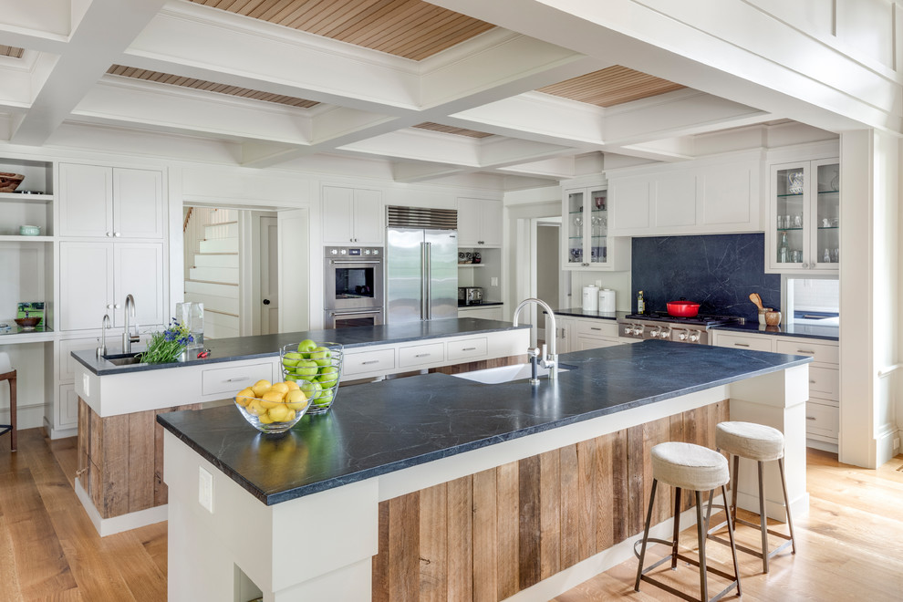 Photo of a large traditional galley open plan kitchen in Boston with a farmhouse sink, beaded inset cabinets, white cabinets, soapstone benchtops, black splashback, stone slab splashback, stainless steel appliances, light hardwood floors and multiple islands.