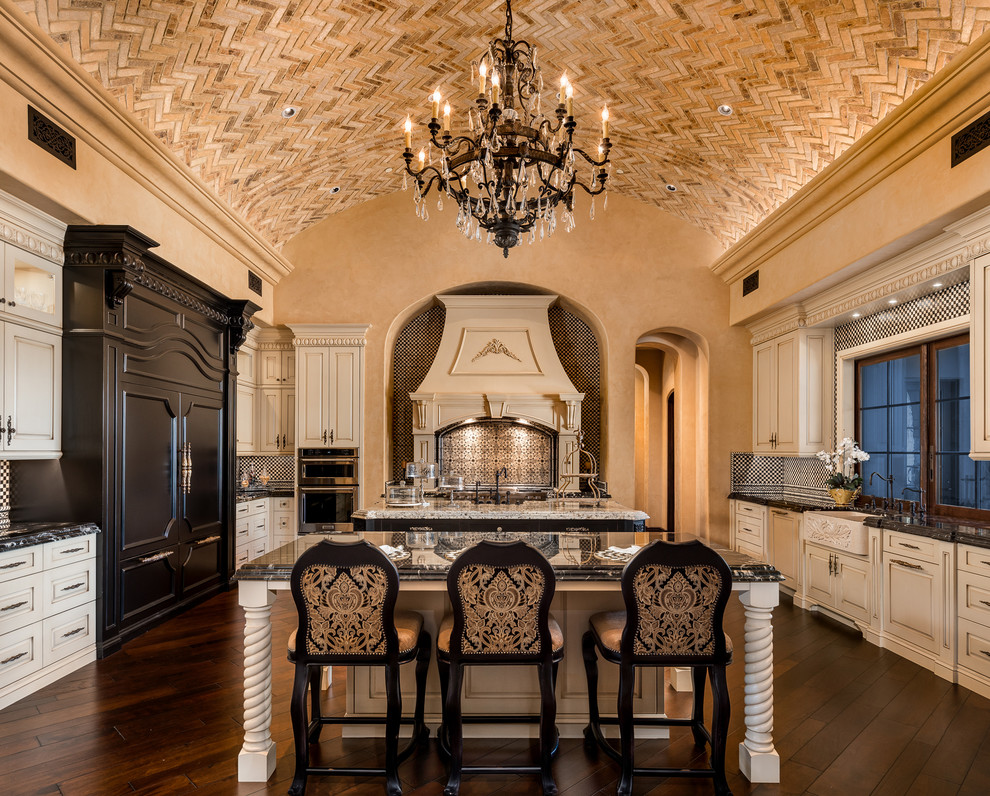 This is an example of an expansive mediterranean u-shaped kitchen in Phoenix with a farmhouse sink, raised-panel cabinets, white cabinets, marble benchtops, beige splashback, terra-cotta splashback, panelled appliances, multiple islands, brown floor, black benchtop and dark hardwood floors.