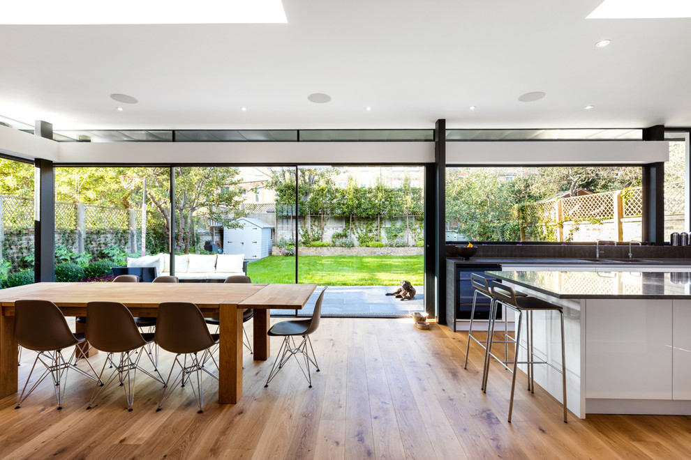 Inspiration for a large contemporary u-shaped eat-in kitchen in London with a drop-in sink, flat-panel cabinets, white cabinets, black splashback, stainless steel appliances, light hardwood floors and with island.
