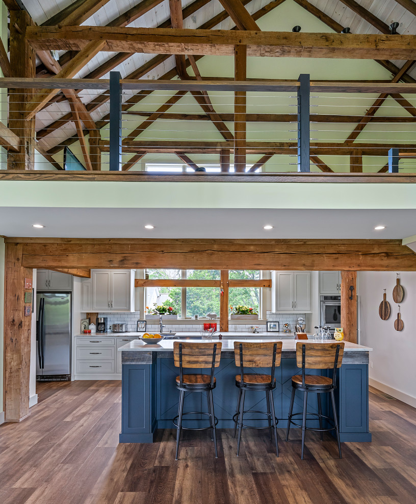 Photo of a mid-sized country galley open plan kitchen in Philadelphia with a farmhouse sink, shaker cabinets, blue cabinets, quartz benchtops, white splashback, porcelain splashback, stainless steel appliances, medium hardwood floors, with island, brown floor, white benchtop and exposed beam.