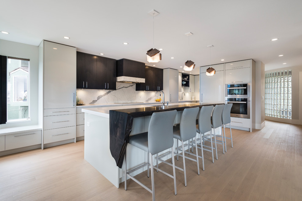 This is an example of a large contemporary l-shaped open plan kitchen in Vancouver with an undermount sink, flat-panel cabinets, white cabinets, quartz benchtops, white splashback, stone slab splashback, panelled appliances, light hardwood floors, with island and brown floor.