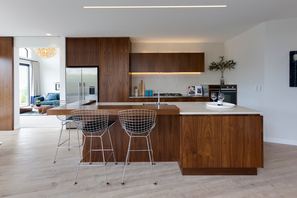 Large contemporary galley eat-in kitchen in Auckland with marble benchtops, white splashback, stainless steel appliances, light hardwood floors, with island, flat-panel cabinets and dark wood cabinets.
