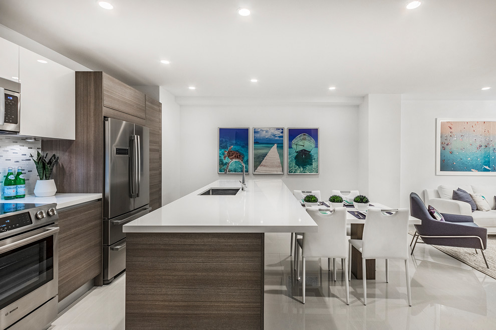 Beach style open plan kitchen in Miami with an undermount sink, flat-panel cabinets, dark wood cabinets, metallic splashback, stainless steel appliances, beige floor and white benchtop.