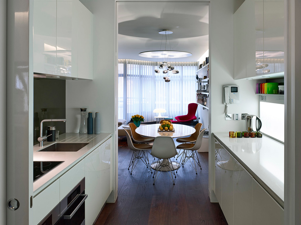This is an example of a small contemporary galley separate kitchen in London with flat-panel cabinets, white cabinets, solid surface benchtops, glass sheet splashback, stainless steel appliances, no island, an undermount sink and dark hardwood floors.