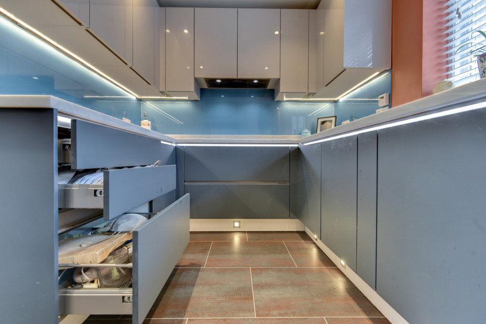 サセックスにある高級な広いモダンスタイルのおしゃれなキッチン (ドロップインシンク、フラットパネル扉のキャビネット、青いキャビネット、人工大理石カウンター、青いキッチンパネル、ガラス板のキッチンパネル、パネルと同色の調理設備、クッションフロア、アイランドなし、茶色い床、白いキッチンカウンター) の写真