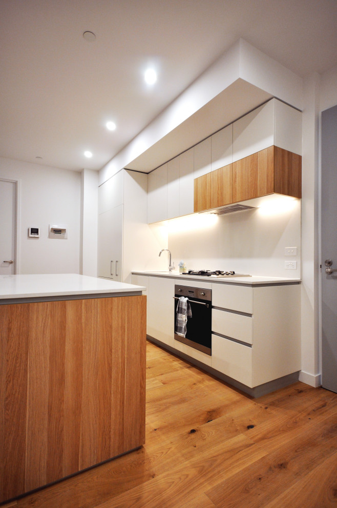 Mid-sized modern single-wall open plan kitchen in Melbourne with an undermount sink, recessed-panel cabinets, medium wood cabinets, solid surface benchtops, white splashback, stainless steel appliances, medium hardwood floors, with island and white benchtop.