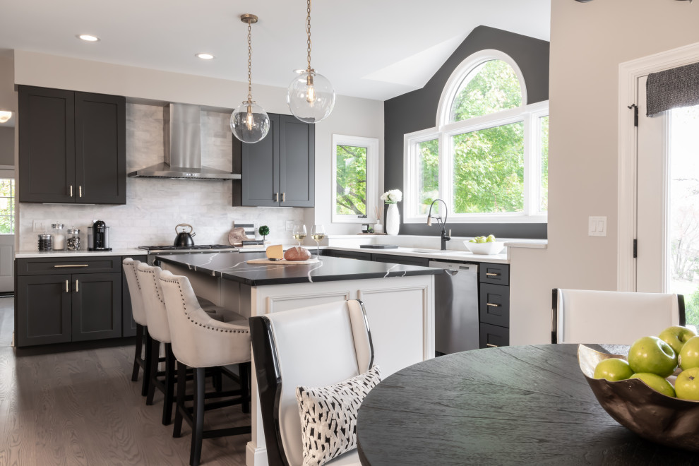 This is an example of a transitional l-shaped eat-in kitchen in St Louis with an undermount sink, shaker cabinets, grey cabinets, quartz benchtops, white splashback, stainless steel appliances, medium hardwood floors, with island, brown floor and black benchtop.
