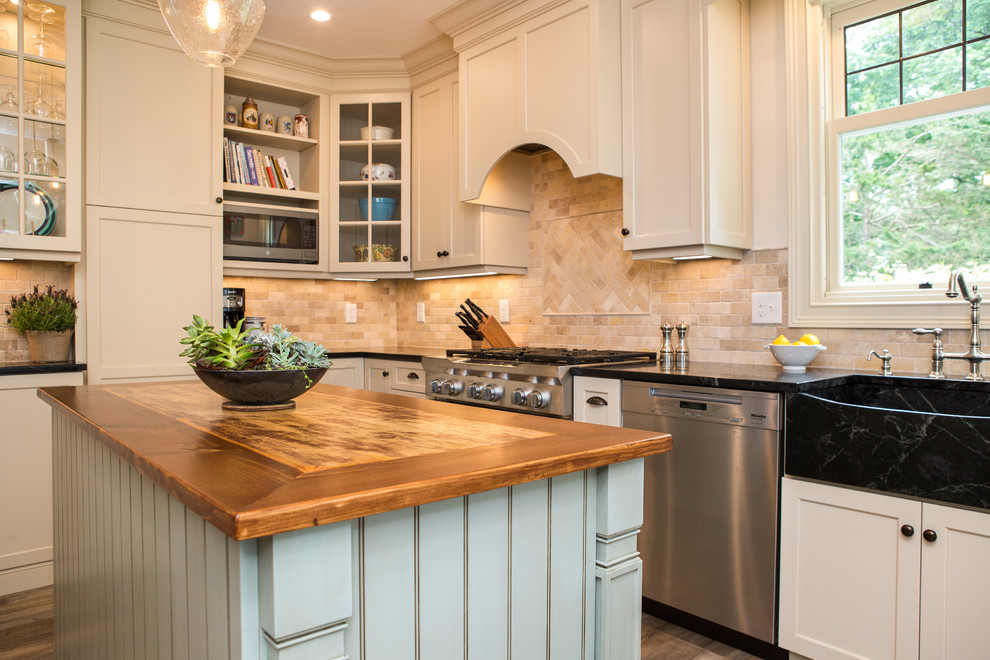 Design ideas for a mid-sized traditional separate kitchen in New York with a farmhouse sink, recessed-panel cabinets, beige cabinets, soapstone benchtops, beige splashback, travertine splashback, stainless steel appliances, vinyl floors, with island, brown floor and black benchtop.