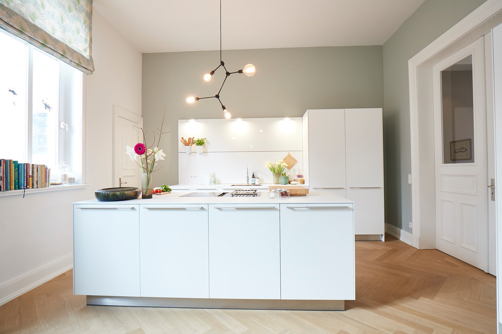 Photo of a mid-sized scandinavian galley separate kitchen in Hamburg with a drop-in sink, flat-panel cabinets, white cabinets, white splashback, panelled appliances, medium hardwood floors, with island, brown floor, white benchtop and glass sheet splashback.