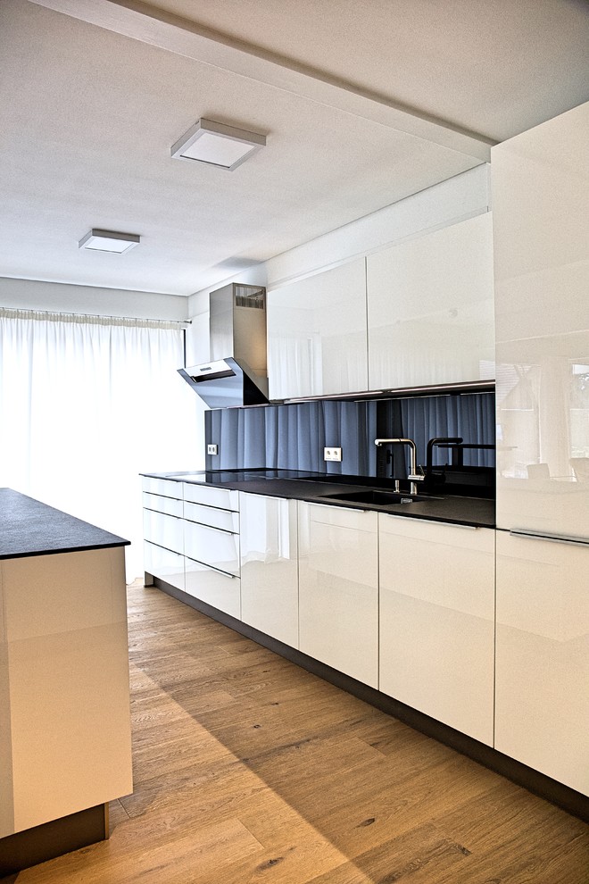 Design ideas for a large contemporary l-shaped open plan kitchen in Berlin with white cabinets, solid surface benchtops, black splashback, glass sheet splashback, with island, flat-panel cabinets, black appliances, light hardwood floors, beige floor and black benchtop.
