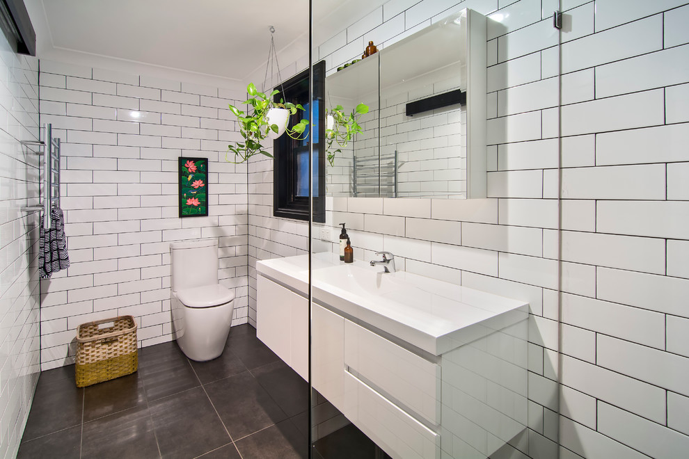 Photo of a small tropical bathroom in Sydney with glass-front cabinets, white cabinets, a curbless shower, a one-piece toilet, white tile, subway tile, white walls, ceramic floors, an integrated sink, solid surface benchtops, black floor, an open shower and white benchtops.