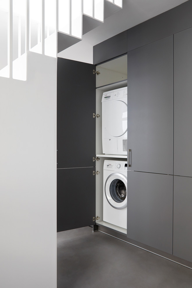 Photo of a contemporary laundry cupboard in London with flat-panel cabinets, grey cabinets, a stacked washer and dryer, grey floor and concrete floors.