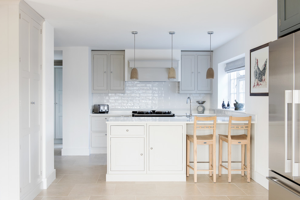 Photo of a mid-sized transitional galley open plan kitchen in Devon with shaker cabinets, grey cabinets, solid surface benchtops, ceramic splashback, stainless steel appliances, limestone floors, a peninsula, a drop-in sink and grey splashback.