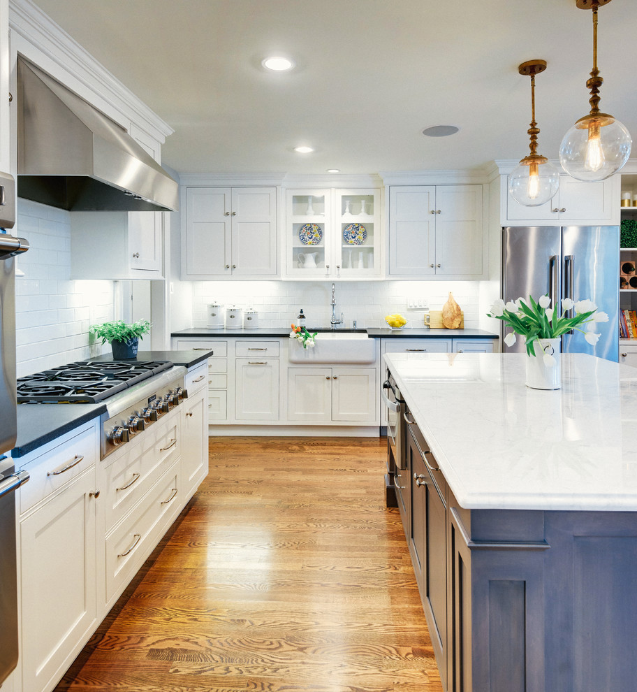 Photo of a transitional l-shaped kitchen in Philadelphia with a farmhouse sink, recessed-panel cabinets, white cabinets, white splashback, subway tile splashback, stainless steel appliances, medium hardwood floors and with island.