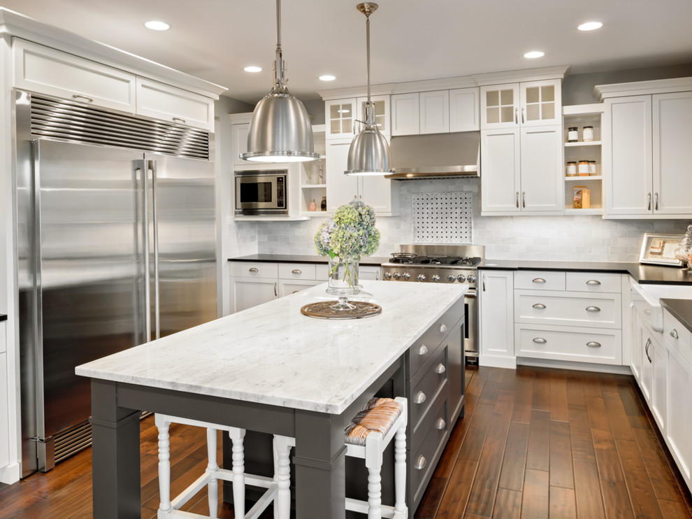 Transitional u-shaped dark wood floor and brown floor kitchen photo in Orange County with a farmhouse sink, shaker cabinets, white cabinets, white backsplash, subway tile backsplash, stainless steel appliances, an island and black countertops
