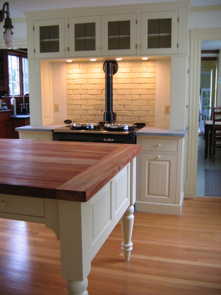 This is an example of a country eat-in kitchen in Portland Maine with raised-panel cabinets, white cabinets, soapstone benchtops, white splashback, black appliances, light hardwood floors and with island.