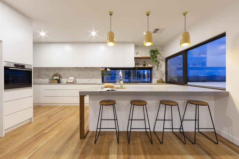 Photo of a mid-sized contemporary l-shaped kitchen in Canberra - Queanbeyan with ceramic splashback, medium hardwood floors, an undermount sink, flat-panel cabinets, white cabinets, grey splashback, black appliances, brown floor and grey benchtop.