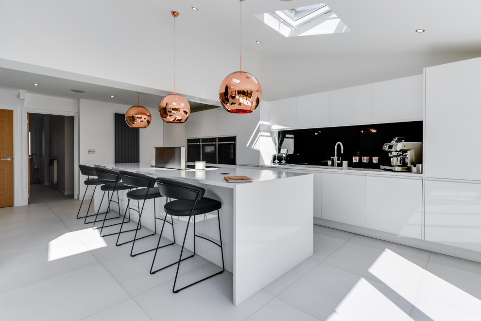 This is an example of a large contemporary single-wall kitchen/diner in Manchester with a submerged sink, flat-panel cabinets, white cabinets, black splashback, black appliances, an island, white floors, white worktops and feature lighting.