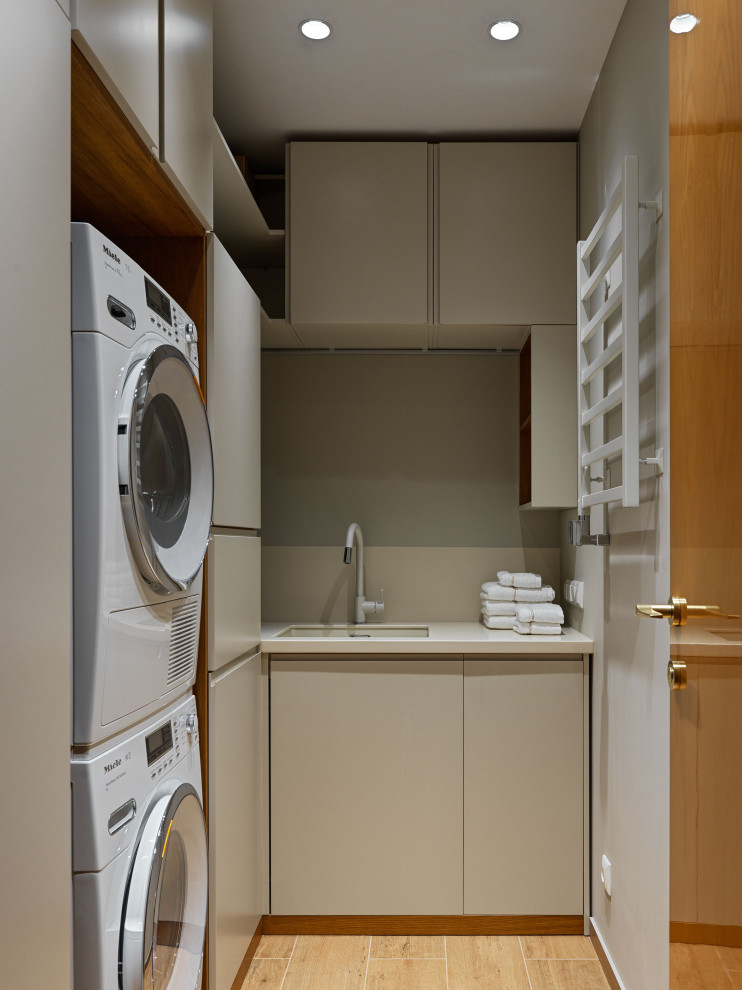 Contemporary l-shaped dedicated laundry room in Moscow with an undermount sink, flat-panel cabinets, beige cabinets, a stacked washer and dryer, brown floor and white benchtop.