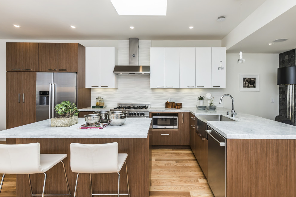 Example of a trendy l-shaped light wood floor and beige floor kitchen design in Other with a farmhouse sink, flat-panel cabinets, medium tone wood cabinets, white backsplash, stainless steel appliances, an island and white countertops