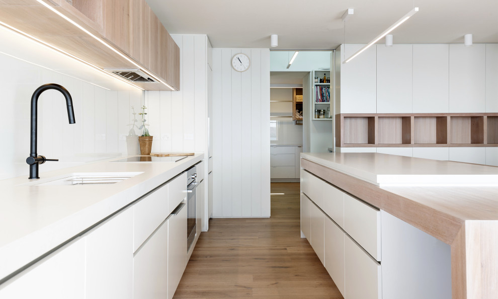 На фото: параллельная кухня-гостиная среднего размера в скандинавском стиле с плоскими фасадами, белыми фасадами, столешницей из кварцевого агломерата, белым фартуком, фартуком из стекла, светлым паркетным полом, островом и белой столешницей с