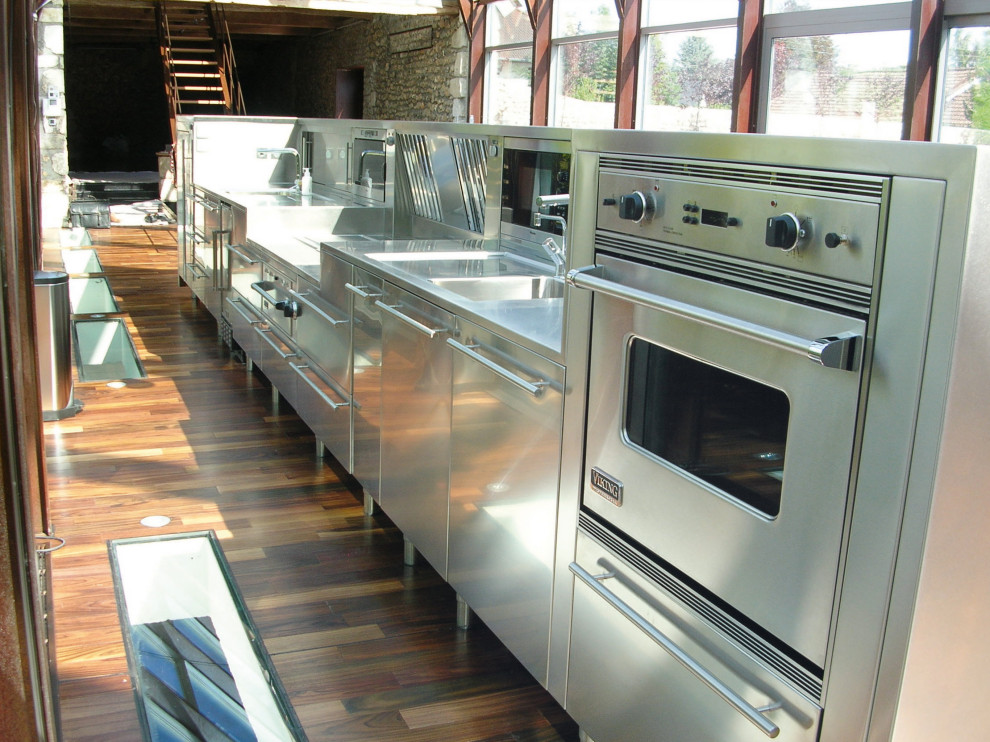 Esempio di un'ampia cucina industriale con lavello integrato, ante in acciaio inossidabile, top in acciaio inossidabile, elettrodomestici in acciaio inossidabile, pavimento in legno massello medio, pavimento marrone e top grigio