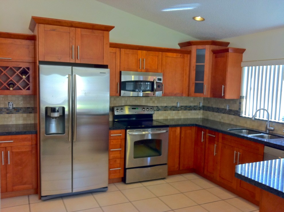 Large transitional l-shaped porcelain tile and beige floor enclosed kitchen photo with a double-bowl sink, shaker cabinets, medium tone wood cabinets, granite countertops, beige backsplash, porcelain backsplash, stainless steel appliances, a peninsula and black countertops