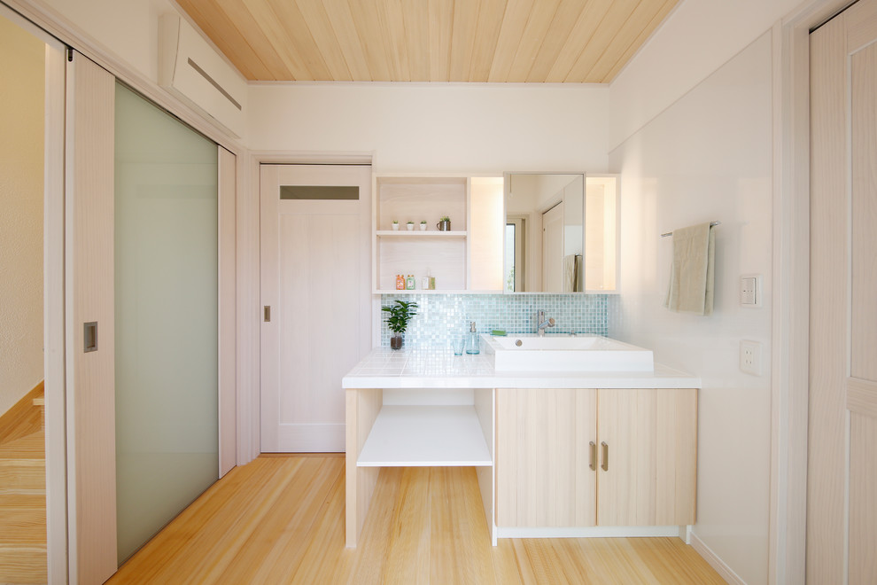 Mid-sized contemporary powder room in Other with beaded inset cabinets, beige cabinets, blue tile, glass tile, white walls, light hardwood floors, a vessel sink, tile benchtops, beige floor and white benchtops.