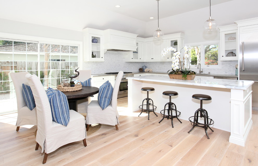 Photo of a beach style l-shaped eat-in kitchen in Orange County with white cabinets, white splashback, stainless steel appliances, light hardwood floors and beige floor.