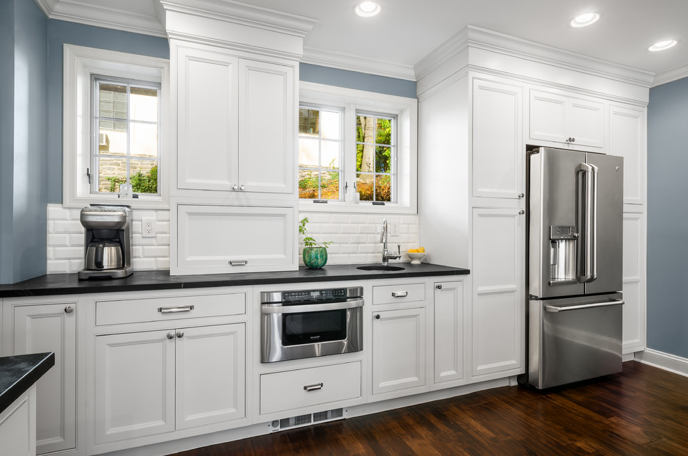 This is an example of a large traditional l-shaped eat-in kitchen in Philadelphia with a farmhouse sink, recessed-panel cabinets, white cabinets, soapstone benchtops, white splashback, subway tile splashback, stainless steel appliances, dark hardwood floors and with island.