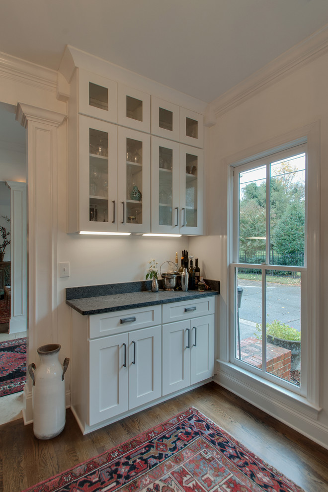 This is an example of a small traditional single-wall home bar in Nashville with shaker cabinets, white cabinets, granite benchtops, medium hardwood floors, brown floor and black benchtop.
