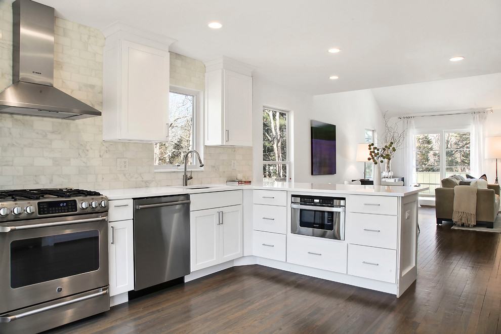 Photo of a transitional l-shaped open plan kitchen in New York with an undermount sink, flat-panel cabinets, white cabinets, white splashback, subway tile splashback, stainless steel appliances, dark hardwood floors, a peninsula, solid surface benchtops, brown floor and white benchtop.