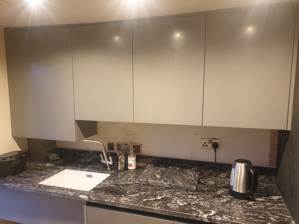 Mid-sized contemporary l-shaped laundry room in West Midlands with flat-panel cabinets, granite benchtops, black splashback, engineered quartz splashback, black benchtop, a drop-in sink, black cabinets, travertine floors, beige floor and vaulted.