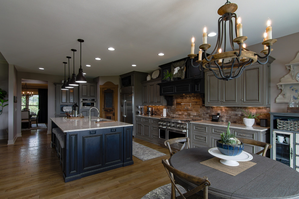 Large mountain style l-shaped medium tone wood floor and brown floor eat-in kitchen photo with an undermount sink, raised-panel cabinets, gray cabinets, multicolored backsplash, stone tile backsplash, stainless steel appliances, an island, beige countertops and granite countertops