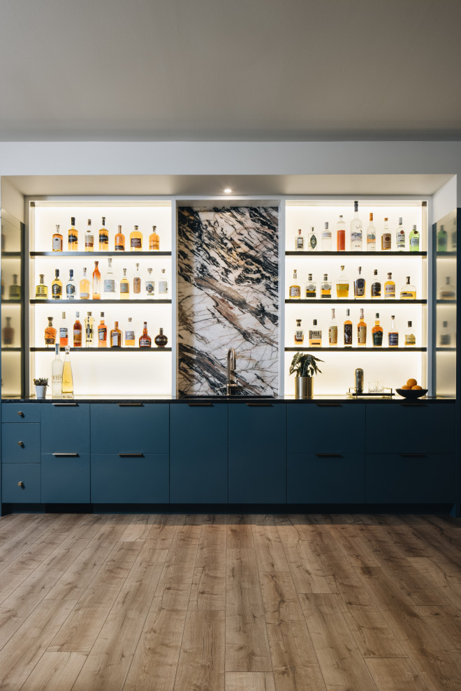 This is an example of a large transitional single-wall wet bar in Chicago with a drop-in sink, flat-panel cabinets, blue cabinets, multi-coloured splashback, light hardwood floors, beige floor and black benchtop.
