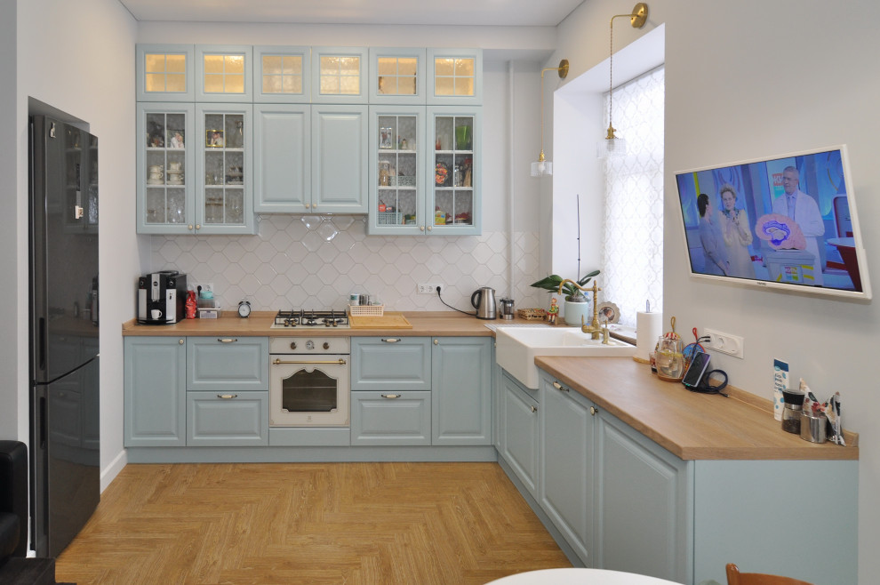 This is an example of a medium sized mediterranean l-shaped kitchen/diner in Other with a belfast sink, raised-panel cabinets, blue cabinets, wood worktops, white splashback, ceramic splashback, white appliances, medium hardwood flooring, no island, beige floors, beige worktops and a drop ceiling.