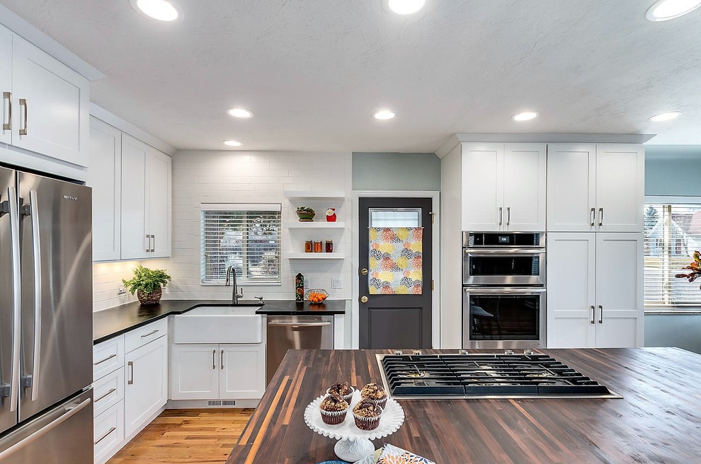 This is an example of a mid-sized contemporary l-shaped kitchen in Denver with a farmhouse sink, shaker cabinets, white cabinets, wood benchtops, white splashback, ceramic splashback, stainless steel appliances, light hardwood floors, with island, beige floor and black benchtop.