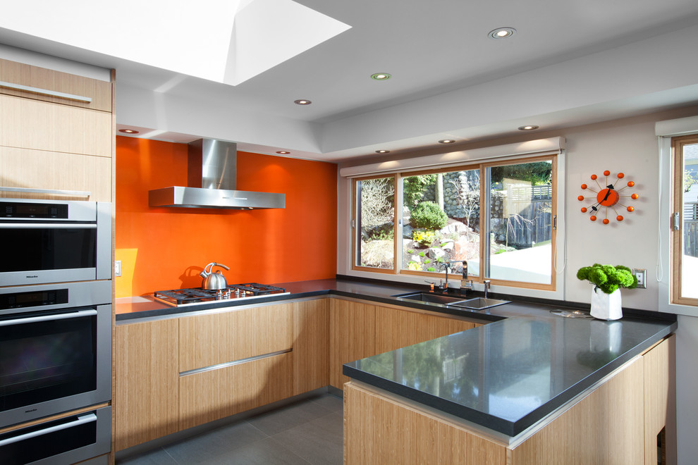 Photo of a mid-sized midcentury u-shaped eat-in kitchen in Vancouver with flat-panel cabinets, light wood cabinets, concrete benchtops, orange splashback, glass sheet splashback, stainless steel appliances, an undermount sink, ceramic floors, a peninsula and grey floor.