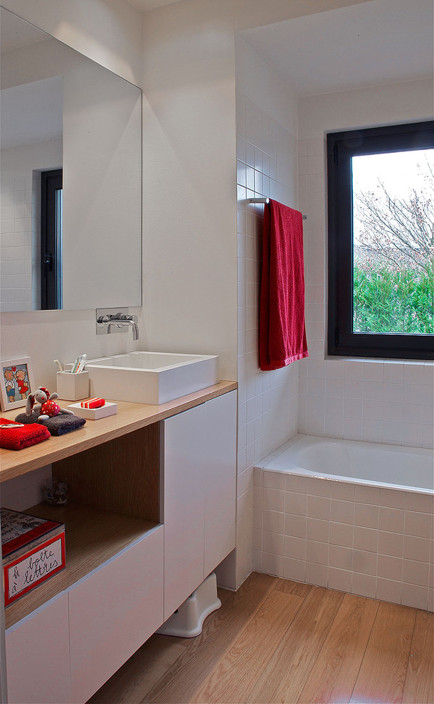 Ejemplo de cuarto de baño infantil moderno de tamaño medio con armarios con paneles lisos, puertas de armario de madera clara, bañera empotrada, baldosas y/o azulejos blancos, baldosas y/o azulejos de cerámica, paredes blancas, suelo de madera clara, lavabo sobreencimera y encimera de madera