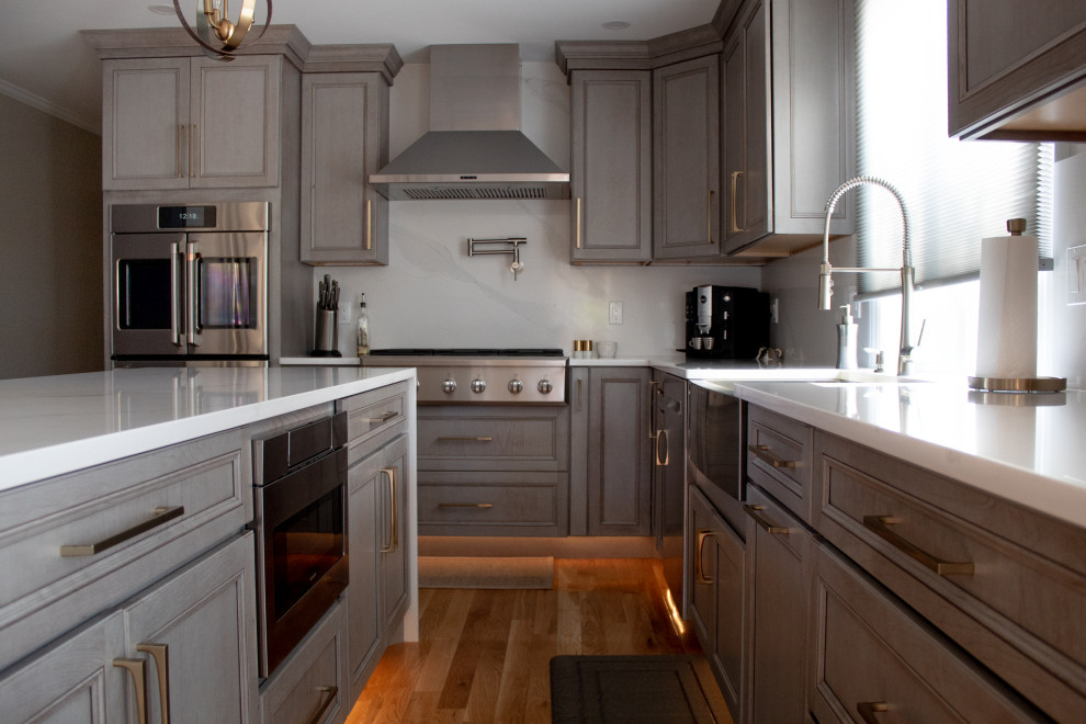 ボストンにある高級な広いコンテンポラリースタイルのおしゃれなキッチン (エプロンフロントシンク、フラットパネル扉のキャビネット、グレーのキャビネット、珪岩カウンター、白いキッチンパネル、クオーツストーンのキッチンパネル、シルバーの調理設備、淡色無垢フローリング、茶色い床、白いキッチンカウンター) の写真