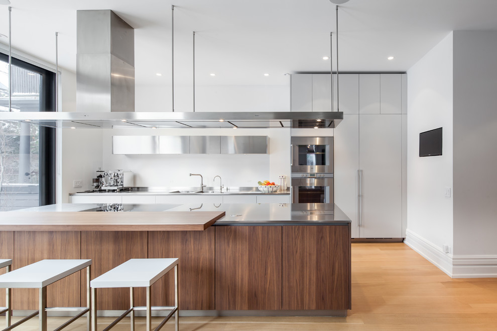 Contemporary galley kitchen in Toronto with an integrated sink, flat-panel cabinets, stainless steel cabinets, stainless steel benchtops, stainless steel appliances, light hardwood floors and with island.