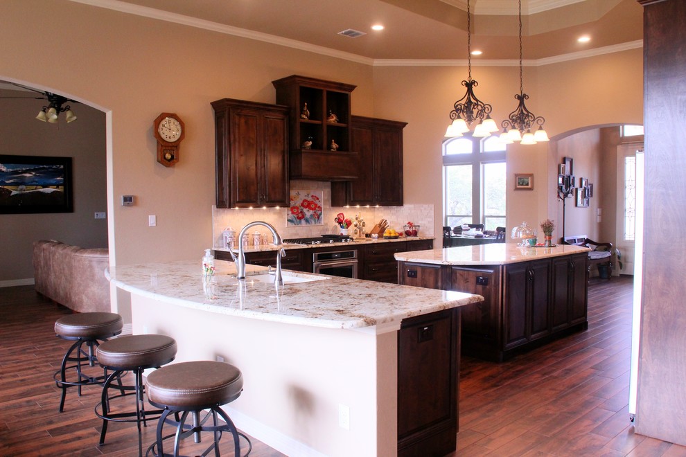 Inspiration for a mid-sized rustic u-shaped dark wood floor and brown floor enclosed kitchen remodel in Austin with an island, an undermount sink, raised-panel cabinets, dark wood cabinets, granite countertops, beige backsplash, stone tile backsplash and stainless steel appliances