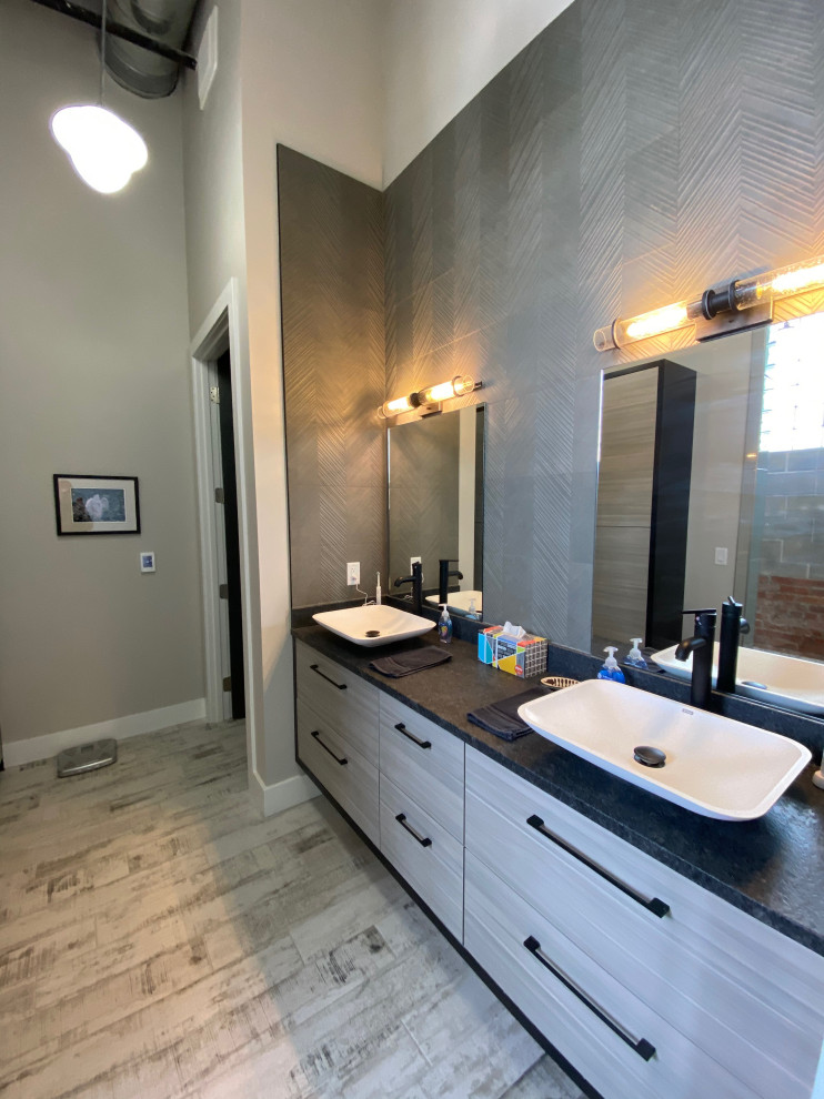 Design ideas for a large industrial master wet room bathroom in Kansas City with flat-panel cabinets, grey cabinets, gray tile, porcelain tile, grey walls, concrete floors, a vessel sink, granite benchtops, grey floor, an open shower, black benchtops, a double vanity, a built-in vanity, exposed beam and brick walls.