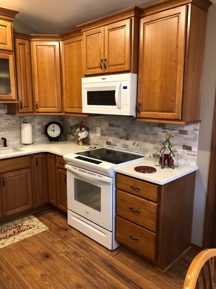 Design ideas for a mid-sized transitional u-shaped eat-in kitchen in Omaha with an undermount sink, raised-panel cabinets, medium wood cabinets, quartz benchtops, beige splashback, stone tile splashback, white appliances, laminate floors, brown floor and white benchtop.