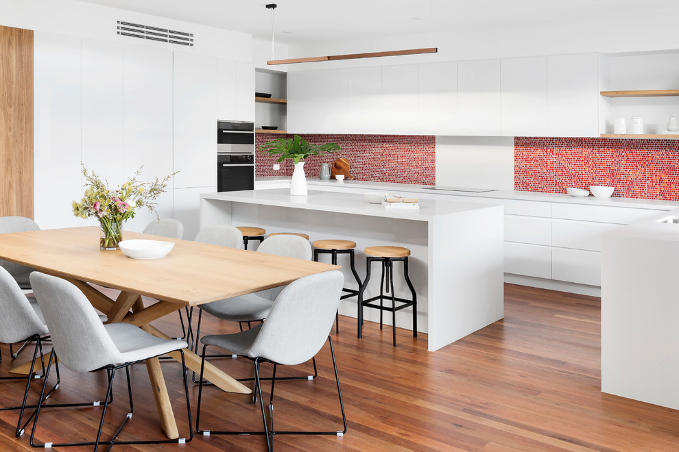 This is an example of a contemporary l-shaped kitchen in Melbourne with an undermount sink, flat-panel cabinets, white cabinets, red splashback, mosaic tile splashback, dark hardwood floors, with island, brown floor and white benchtop.